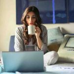 woman-in-grey-jacket sits on bed uses grey laptop