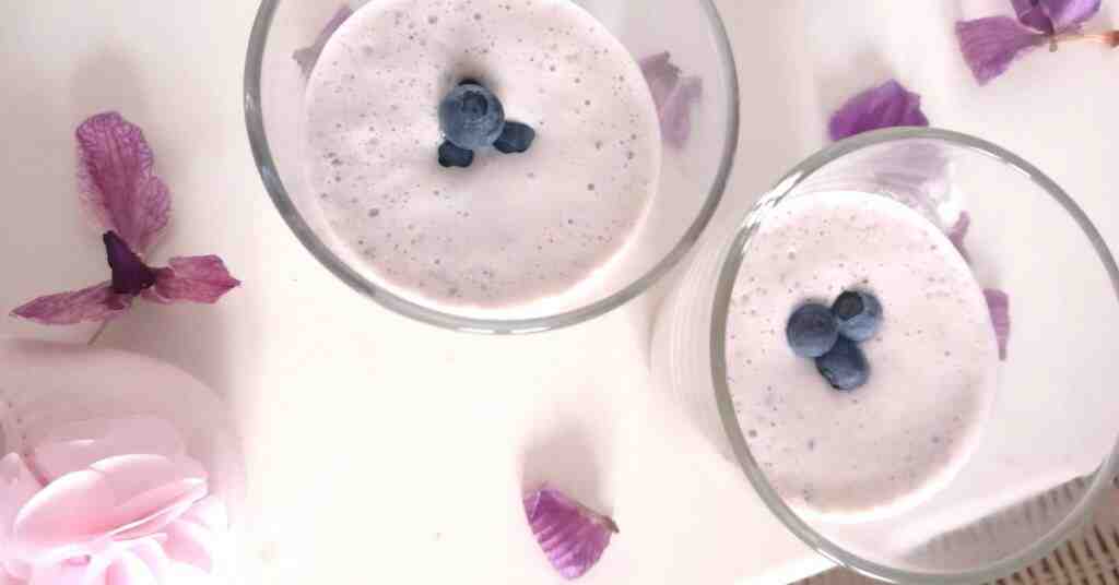 Two berry smoothies with blueberries on top and paleonopsis petals spread on the table