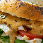 homemade sweet onion sauce, glossy and golden, drizzling over a fresh Subway-style sandwich. The sauce has a smooth, velvety texture with tiny specks of onion flakes and poppy seeds. Shot on a marble countertop with natural lighting, fresh lettuce, tomatoes, and turkey visible in the sandwich.